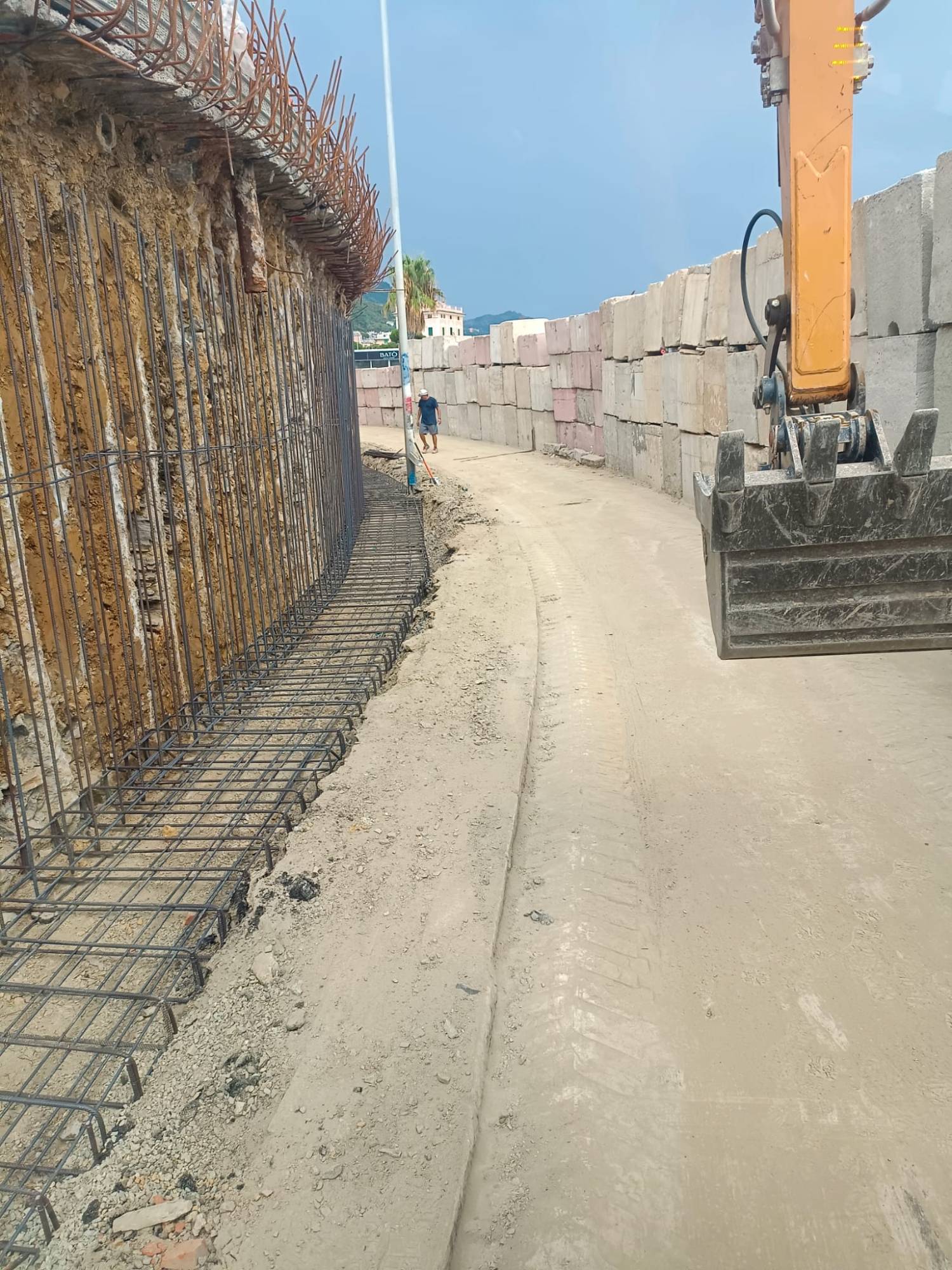 Armatura in ferro di un muro contro il fronte di scavo in roccia, con una fila di blocchi di cemento sull’altro lato della strada