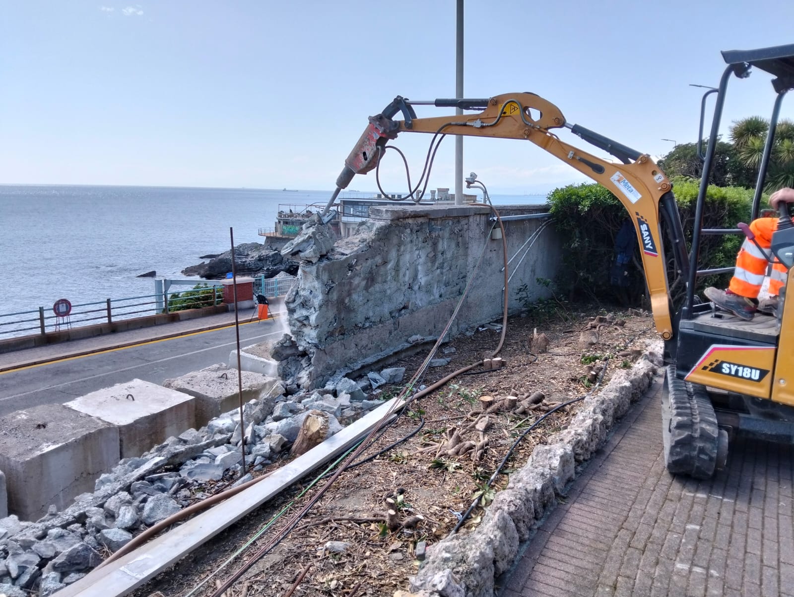 Un miniescavatore con il martello demolitore abbatte un vecchio muro affacciato sul mare