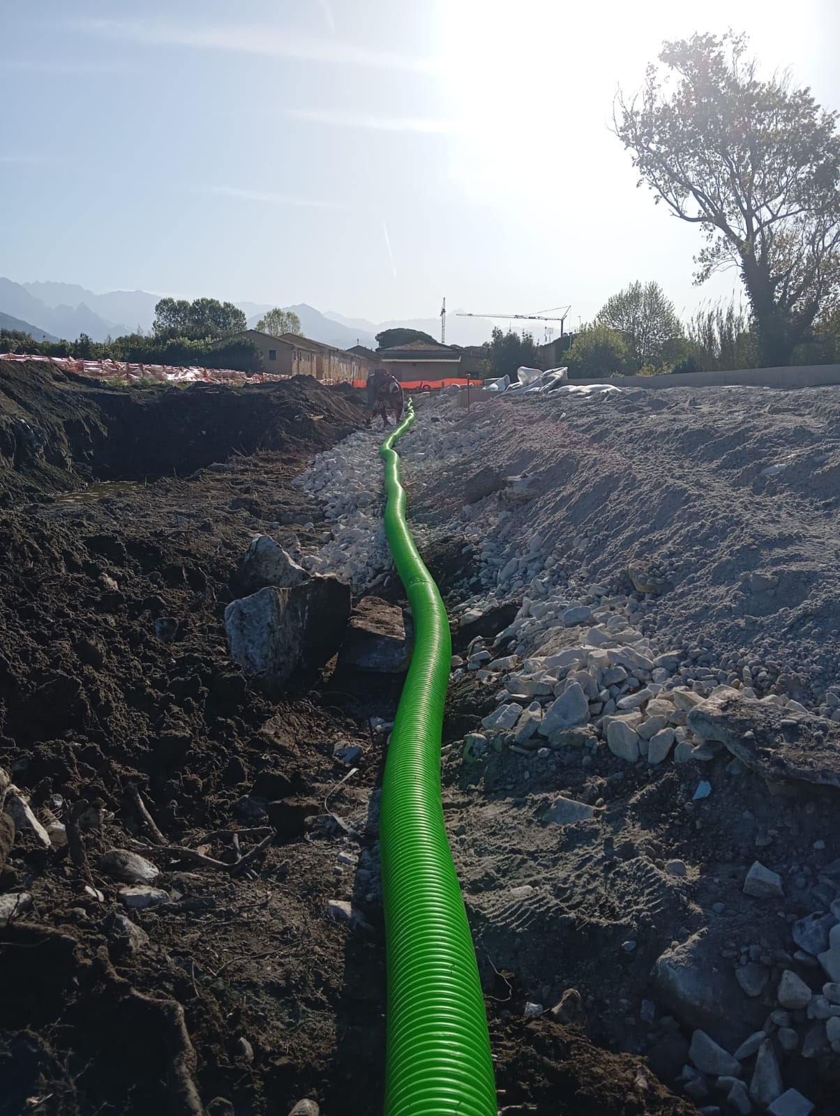 Tubo drenante verde nella trincea fra la terra scavata e il pietrame, con un operaio in fondo