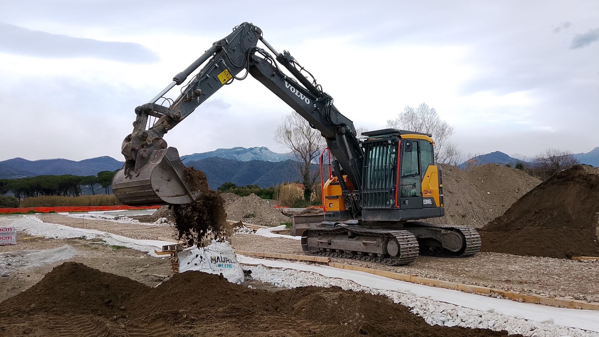 Un escavatore scarica terra accanto al sottofondo di una strada, fra il geotessile e il pietrame, con le montagne sullo sfondo