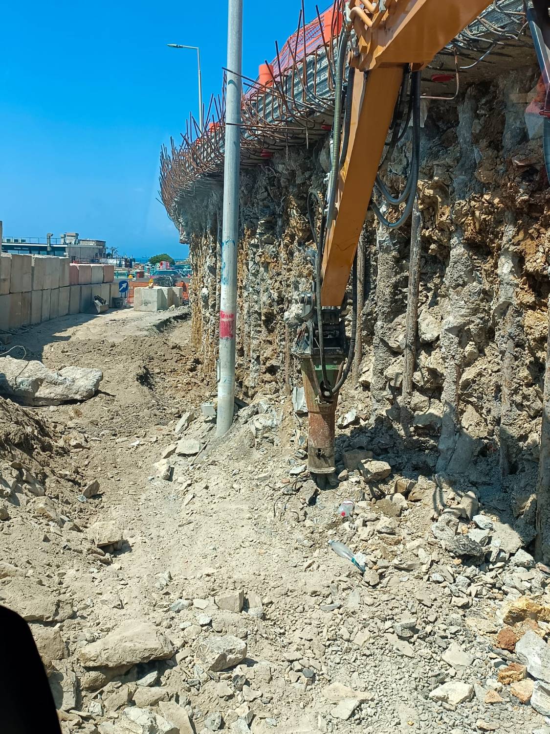 Il martello demolitore dell’escavatore al lavoro al piede di un fronte di roccia sotto una trave armata