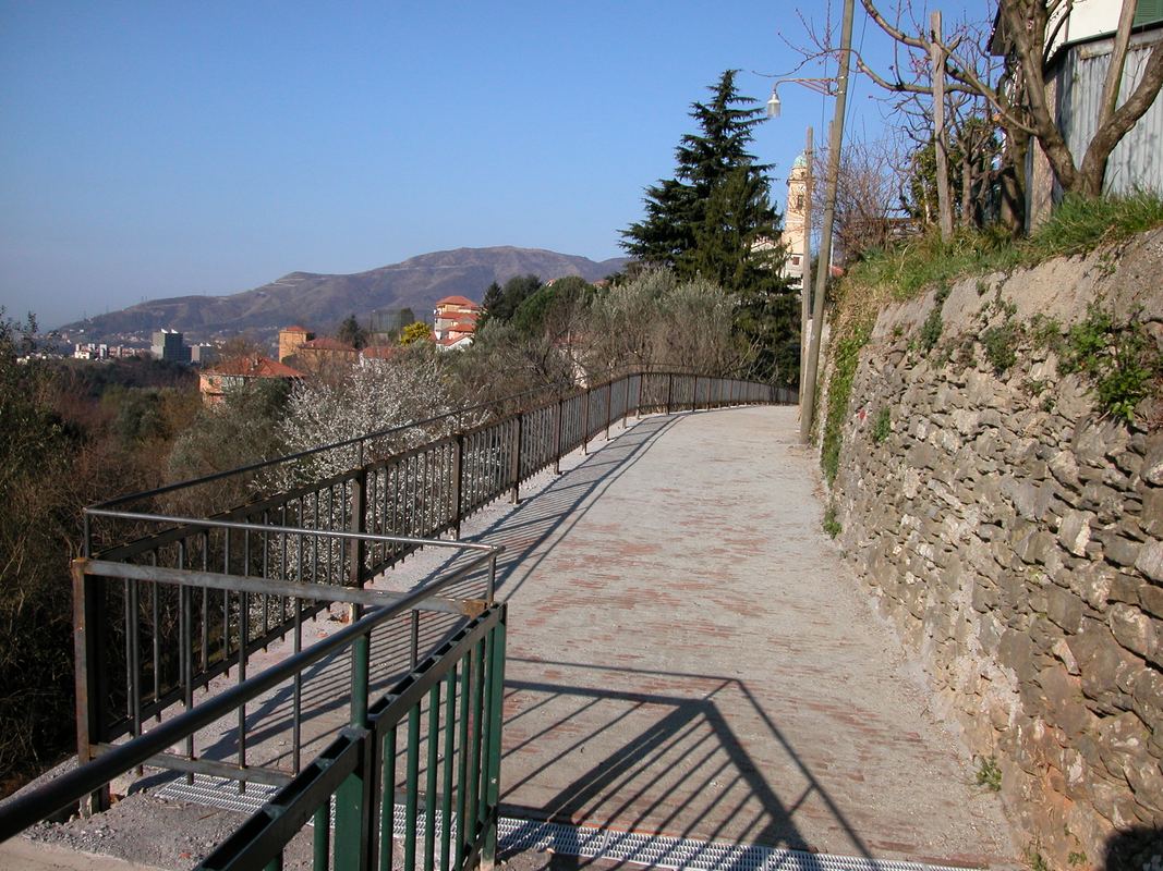 Percorso pedonale pavimentato in mattoni posati di coltello, con ringhiera e vista sulle alture