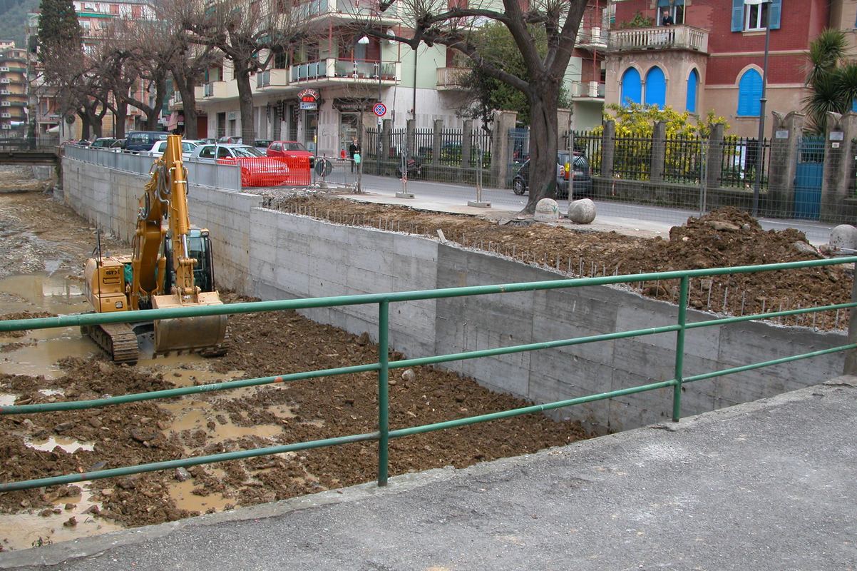 Muro d’argine in cemento appena gettato lungo il torrente, con escavatore e ringhiera