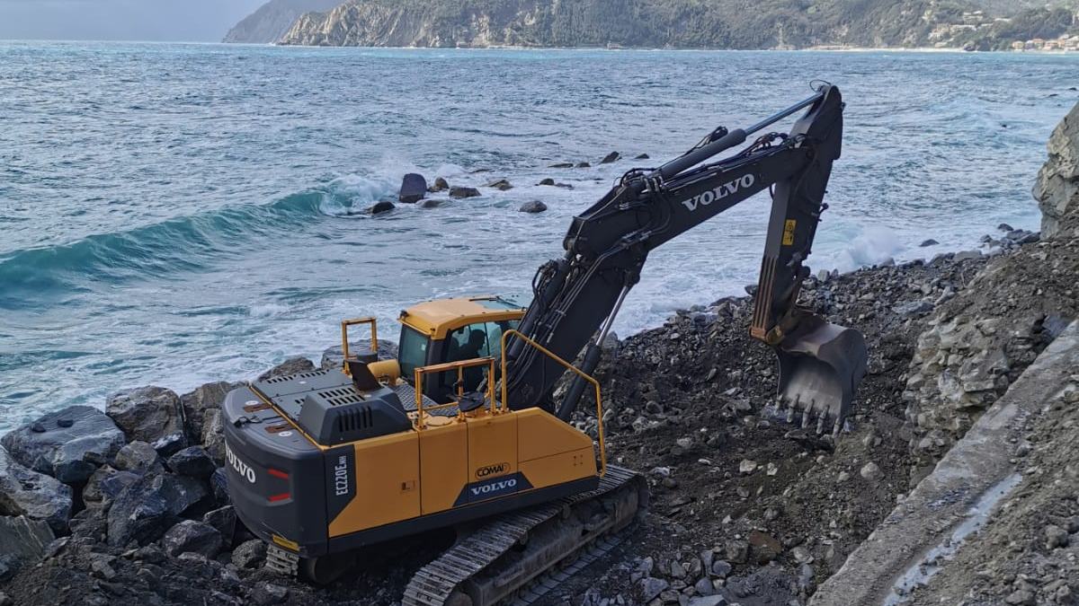 Escavatore cingolato al lavoro sulla scogliera, con la costa del levante sullo sfondo