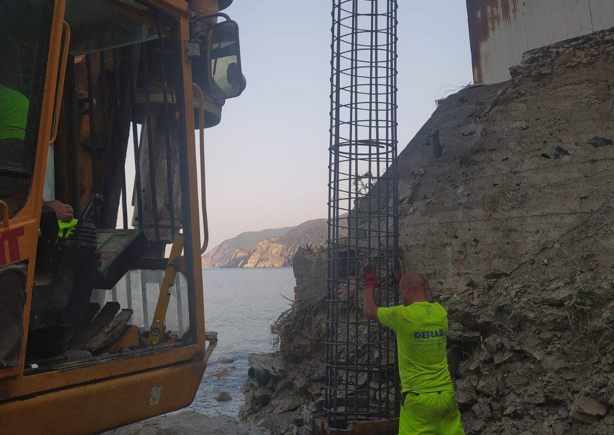 Un operaio guida la gabbia d'armatura nel foro del palo, con la trivella e il mare alle spalle
